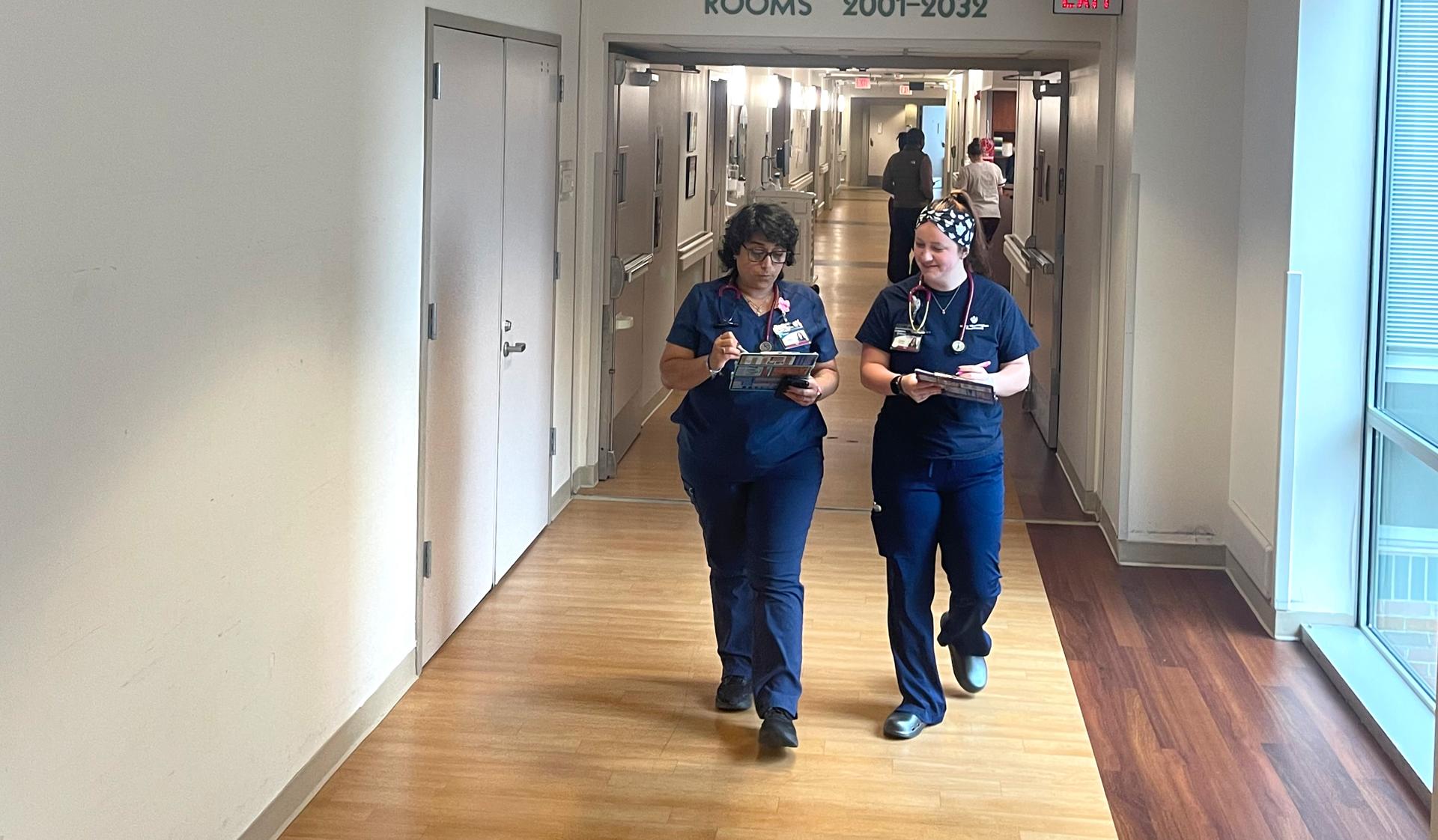 Two critical care nurses having a conversation and walking through corridor with others in the background.