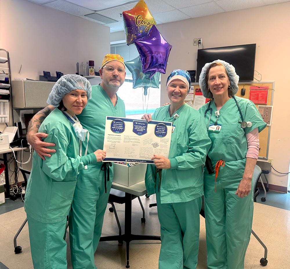 Group of four anesthesiology team members holding up a poster showing three recognition awards in front of congratulatory balloons.