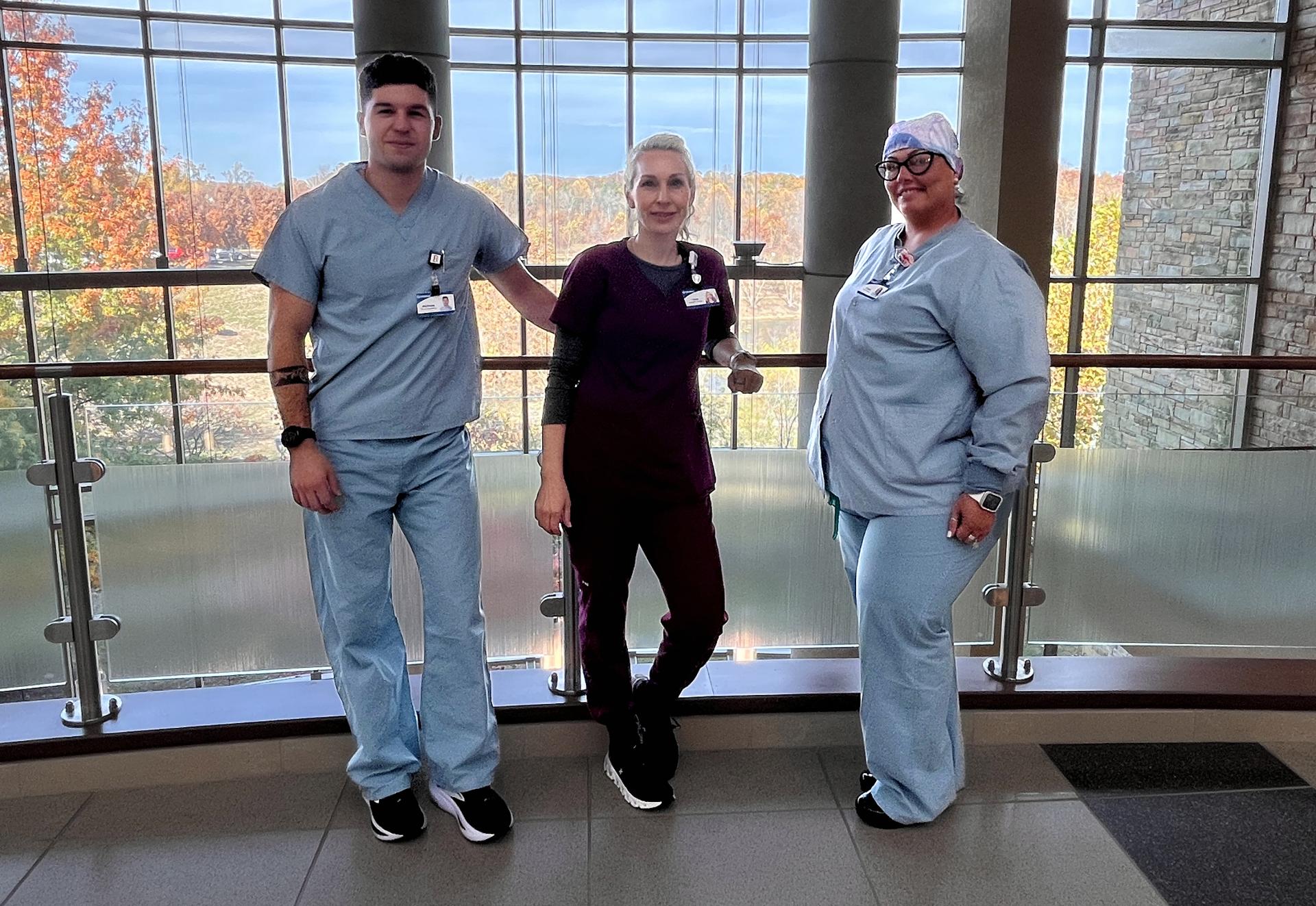 Trio of employees in scrubs smiling and standing in front of picturesque scene of fall foliage.