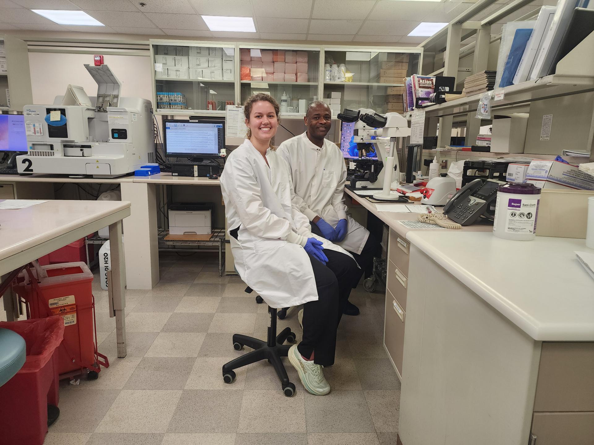 Two smiling medical laboratory employees sitting at a desk surrounded by lab instruments.